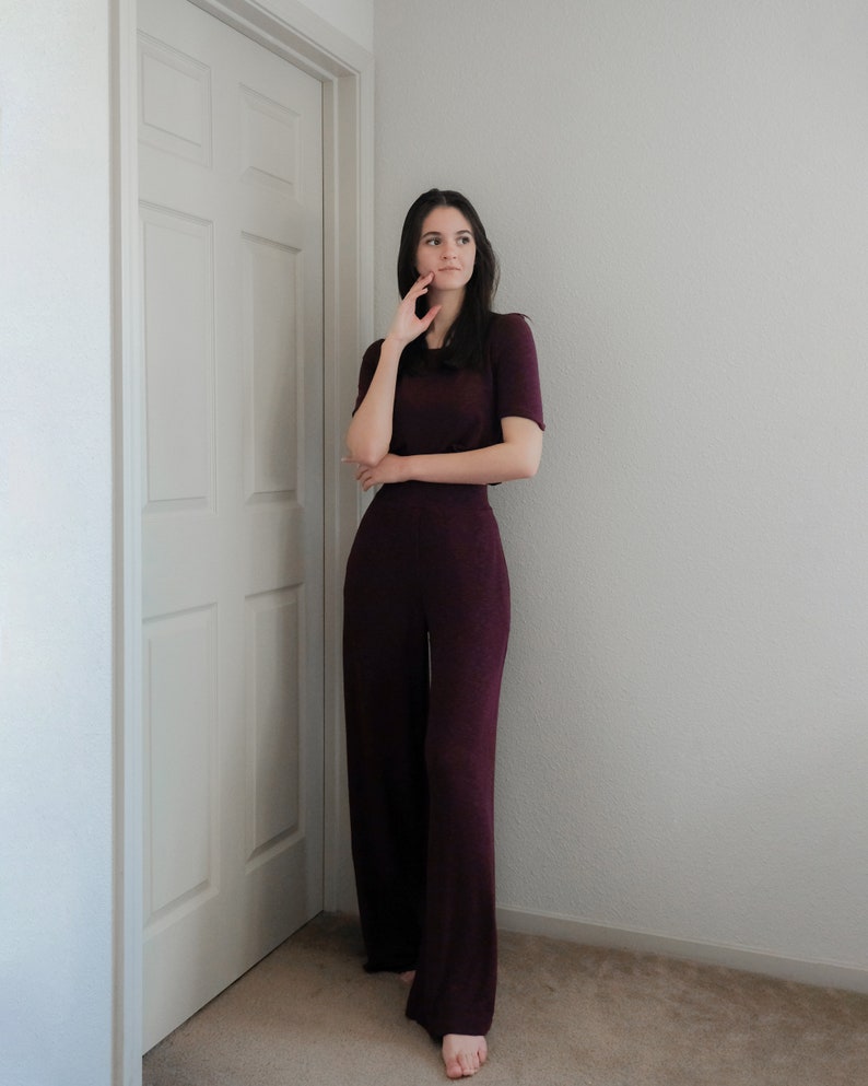 Op de afbeelding: Een bordeauxrode tweedelige outfit bestaande uit een top met korte mouwen en een broek met wijde pijpen. De outfit is gemaakt van een zachte, getextureerde stof. Het model staat in de buurt van een witte deur en een lichtgekleurde muur.