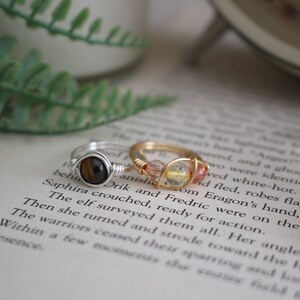 May include: Two wire-wrapped rings with gemstone accents. The ring on the left has a tiger's eye stone, while the ring on the right has a mix of pink and yellow stones.