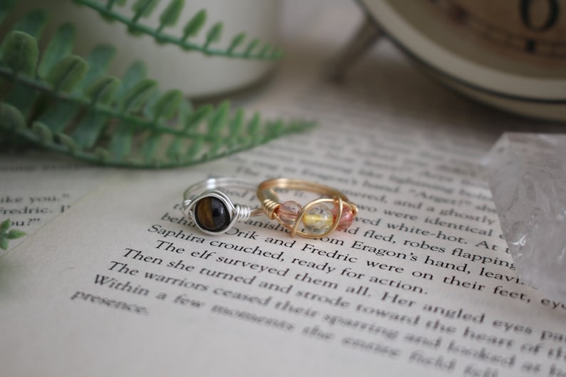 May include: Two wire-wrapped rings with gemstone accents. The ring on the left has a tiger's eye stone, while the ring on the right has a mix of pink and yellow stones.
