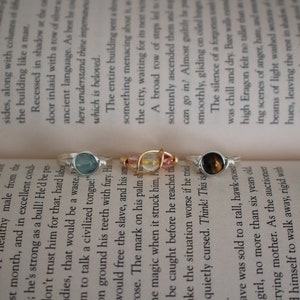 May include: Three silver rings with different colored stones. The rings are on a book with text. The stones are a light blue, a clear stone with gold flecks, and a brown and gold stone.