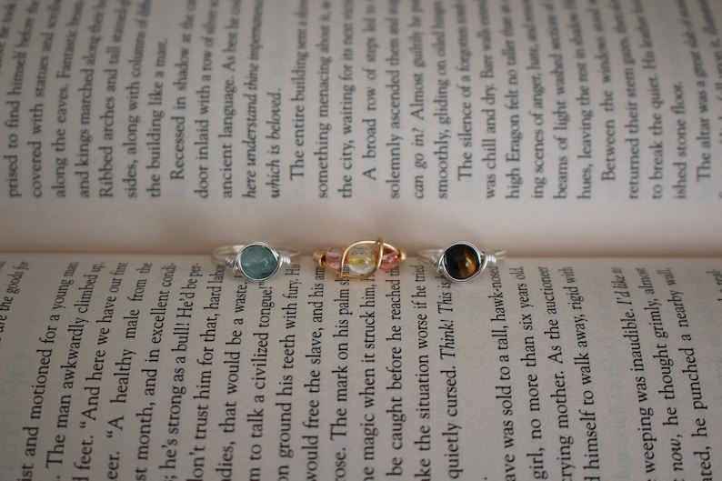 May include: Three silver rings with different colored stones. The rings are on a book with text. The stones are a light blue, a clear stone with gold flecks, and a brown and gold stone.