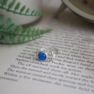 May include: A silver ring with a blue gemstone set in a wire wrapped setting. The ring is on a book with text.