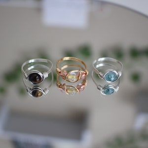 May include: Three silver wire wrapped rings with different stones. The first ring has a brown tiger's eye stone, the second ring has a clear stone with pink and gold accents, and the third ring has a blue-green stone.
