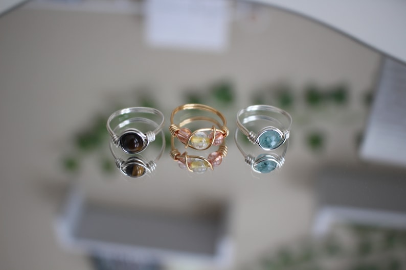 May include: Three silver wire wrapped rings with different stones. The first ring has a brown tiger's eye stone, the second ring has a clear stone with pink and gold accents, and the third ring has a blue-green stone.