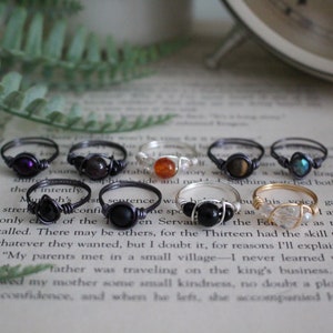 May include: A collection of ten wire-wrapped rings with various gemstone beads. The rings are displayed on a book with a white background. The gemstones include amethyst, tiger's eye, carnelian, black onyx, and clear quartz.
