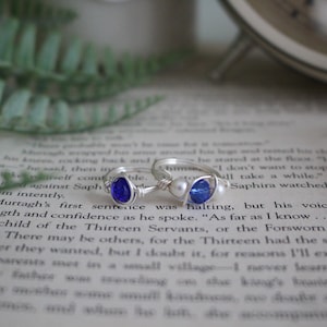 May include: Two silver rings with blue and white glass beads. The ring on the left has a dark blue teardrop-shaped bead. The ring on the right has a white pearl and a light blue round bead.