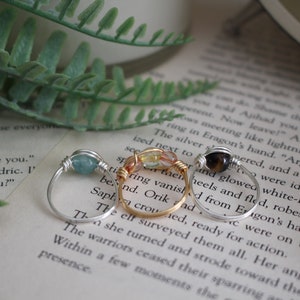 May include: Three silver wire wrapped rings with different colored stones. The first ring has a light blue stone, the second ring has a light green and orange stone, and the third ring has a brown stone.