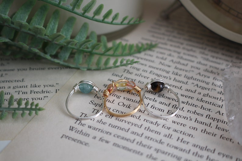 May include: Three silver wire wrapped rings with different colored stones. The first ring has a light blue stone, the second ring has a light green and orange stone, and the third ring has a brown stone.