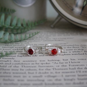 May include: Two silver rings with red gemstone accents. The rings are on a book with text. The rings are simple and elegant.