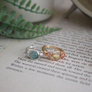 May include: Two wire-wrapped rings with gemstones. The ring on the left has a light blue gemstone and a silver wire. The ring on the right has a pink and yellow gemstone and a gold wire.