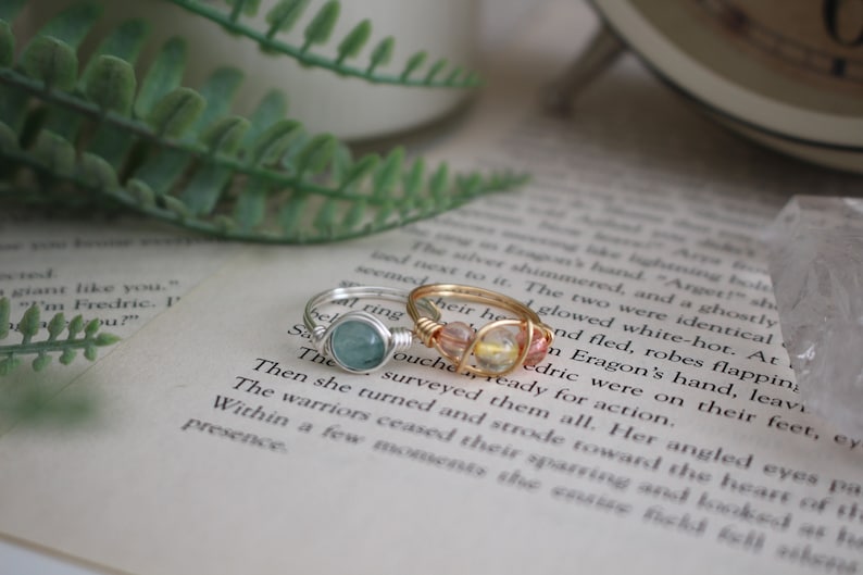 May include: Two wire-wrapped rings with gemstones. The ring on the left has a light blue gemstone and a silver wire. The ring on the right has a pink and yellow gemstone and a gold wire.