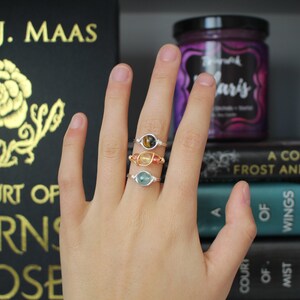May include: Three silver rings with different colored gemstones. The rings are stacked on a finger. The gemstones are a tiger's eye, a carnelian, and an aquamarine.