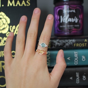 May include: Two rings with gemstones on a hand. The ring on the index finger has a light blue gemstone and a silver band. The ring on the middle finger has a light orange gemstone and a gold band. The hand is in front of a stack of books with the titles "A Court of Frost and Star", "A Court of Wings and Ruin", and "A Court of Mist and Fury".