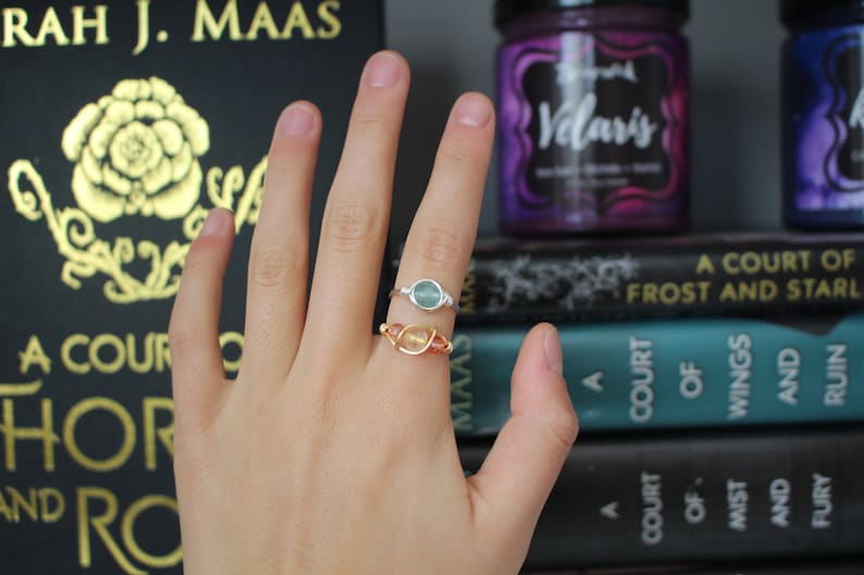 May include: Two rings with gemstones on a hand. The ring on the index finger has a light blue gemstone and a silver band. The ring on the middle finger has a light orange gemstone and a gold band. The hand is in front of a stack of books with the titles "A Court of Frost and Star", "A Court of Wings and Ruin", and "A Court of Mist and Fury".