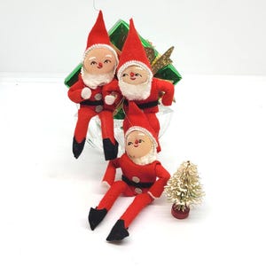 May include: Three vintage Christmas elf ornaments. Each elf is dressed in a red felt suit with a matching hat, black shoes, and a white beard. The elves have painted faces and are posed in a sitting position. A small, snow-covered Christmas tree is also present.