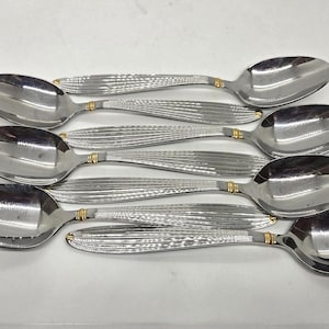May include: A set of five silver-toned stainless steel spoons with a polished finish. Each spoon features a textured handle with vertical ridges and a small gold-colored band accent located where the handle meets the bowl of the spoon.