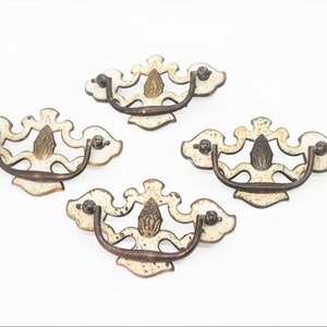 Peut inclure: Quatre poignées de tiroir antiques blanches et dorées avec des motifs ornés. Les poignées ont une poignée en métal brun.