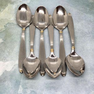 May include: A set of six silver-toned metal spoons arranged in a vertical pattern. Each spoon features a long, slender handle with vertical ribbed detailing and a small gold-colored band accent located near the neck of the spoon bowl.