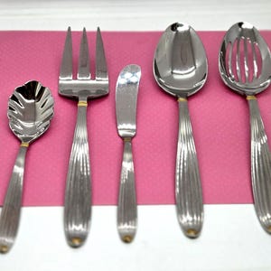 May include: A set of five stainless steel serving utensils featuring ribbed handles with gold-tone accents. The collection includes a scalloped spoon, a three-tined serving fork, a butter knife, a solid serving spoon, and a slotted serving spoon.