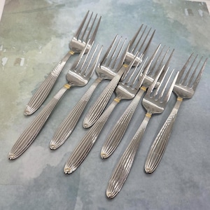 May include: A set of seven silver-toned stainless steel forks featuring a vertical ribbed pattern on the handles and a small gold-colored band accent located just below the tines. Each fork has four tines and a polished finish throughout the metal.