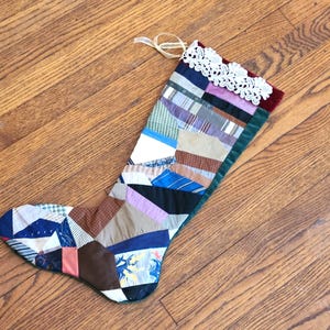 Peut inclure: Une chaussette de Noël en patchwork avec divers motifs de tissus bleus, marron, roses et violets. Le bord supérieur est orné de dentelle blanche et d'une bordure en velours bordeaux. Un ruban crème est attaché pour l'accrocher.