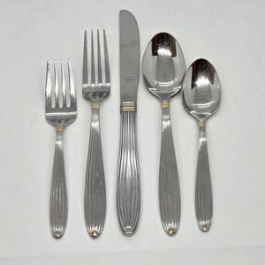 May include: A five-piece stainless steel flatware set consisting of a small fork, a large fork, a dinner knife, a large spoon, and a small spoon. Each piece features a vertical ribbed handle design with a thin gold-tone band near the neck.
