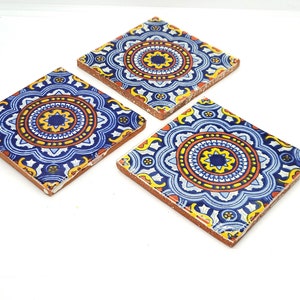 May include: Three square ceramic coasters with a vibrant, intricate design. The coasters feature a blue, yellow, and orange floral pattern on a white background. The edges are a reddish-brown color, and the coasters are arranged on a white surface.