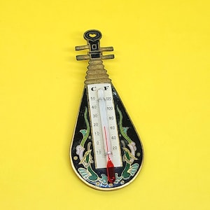 Peut inclure: Un thermomètre noir et or en forme d'instrument de musique avec un liquide rouge à l'intérieur. Le thermomètre a un cadran noir et blanc avec des graduations en Celsius et en Fahrenheit. L'instrument est décoré de motifs floraux verts et blancs.