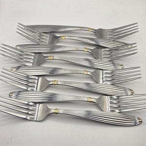 May include: A set of four stainless steel dinner forks arranged in a stack. Each fork features a polished finish with vertical ribbed detailing along the handle and a small gold-toned band accent located near the neck of the utensil.