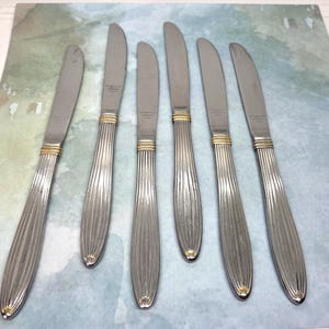 May include: A set of six silver-toned stainless steel dinner knives featuring vertical fluted handles and gold-toned bands near the bolster. Each knife has a smooth, curved blade and a tapered handle design with a rounded end.