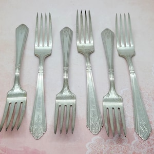 May include: Six silver forks with ornate handles. The forks are arranged in a row on a pink and white background.