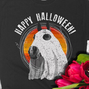 May include: Black t-shirt with a white ghost horse wearing a ghost sheet and the text "Happy Halloween!" printed on it. The shirt is laying on a wooden surface with a bouquet of red tulips and two horseshoes.