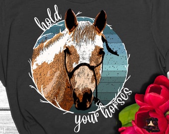 Puede incluir: Una camiseta negra con una ilustración de un caballo blanco y marrón en un círculo con el texto "hold your horses".