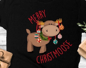 Puede incluir: Camiseta negra con un alce de dibujos animados que lleva una corona y sostiene adornos navideños. El texto "Merry Christmoose" está impreso en la camiseta.