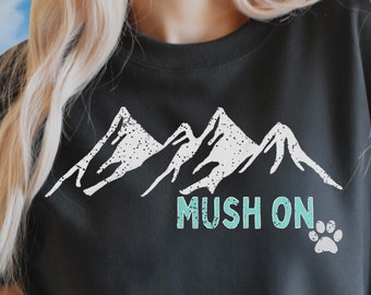 Puede incluir: Sudadera negra con un gráfico de cordillera blanca y el texto "Mush On" en turquesa con una huella de pata.