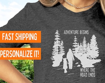 Puede incluir: Una camiseta gris oscura con un gráfico blanco de dos figuras caminando por un bosque. El texto "Adventure Begins Where The Road Ends" está impreso en la camiseta.
