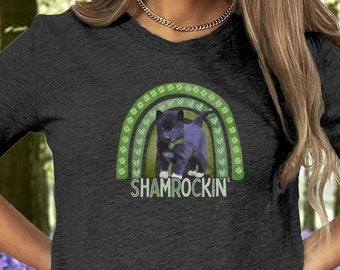 May include: A dark heather grey t-shirt with a green shamrock rainbow and a black cat wearing a green shamrock. The text "Shamrockin'" is printed below the rainbow.