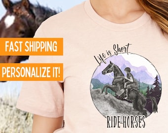 Puede incluir: Una camiseta rosa con una ilustración de acuarela de una persona montando a caballo frente a un paisaje de montaña. El texto "Life is Short Ride Horses" está impreso en la camiseta.
