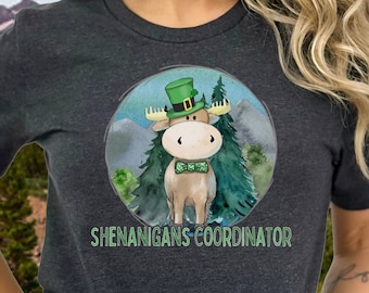 May include: A dark gray short-sleeved t-shirt with a green, white, and brown cartoon moose wearing a green leprechaun hat and a green bow tie. The moose is surrounded by green trees and the text "Shenanigans Coordinator" is printed below the moose.