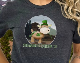 May include: A dark heather grey short-sleeved t-shirt with a graphic of a cartoon moose wearing a green hat and holding a shamrock. The text "Shenanigans Coordinator" is printed below the moose.