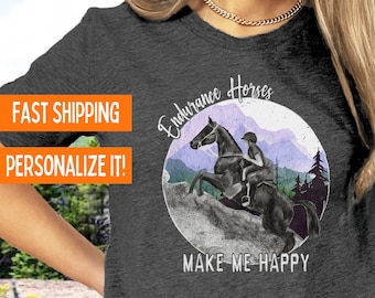 Puede incluir: Camiseta gris de manga corta con una ilustración de acuarela de un jinete a caballo en un paisaje de montaña. El texto "Endurance Horses" y "Make Me Happy" está impreso en la camiseta.