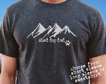 May include: A dark gray short-sleeved t-shirt with a white graphic of mountains and the text "sled dog dad" with a paw print. The text "choose from: short sleeve, long sleeve or sweatshirt!" is printed on the right sleeve. The t-shirt is available in short sleeve or long sleeve and sweatshirt options.  The t-shirt is a Bella Canvas 3001.