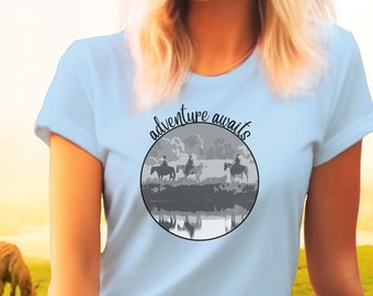 May include: Light blue women's t-shirt with a graphic of three people riding horses in front of a sunset with the text "adventure awaits" in a circle.