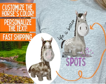 Puede incluir: Una camiseta gris con una ilustración de acuarela de un caballo con manchas. El texto "life is better with spots" está impreso en la camiseta. El color del caballo se puede personalizar.