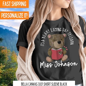 Op de afbeelding: Een zwart T-shirt met een bruine beer die een rood boek draagt en een bos rode rozen vasthoudt. De tekst "It's a Beary Exciting Day to Learn" staat in een cirkel rond de beer. De tekst "Miss Johnson" staat onder de beer.
