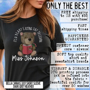 Op de afbeelding: Een zwart T-shirt met een illustratie van een bruine beer die een rood boek vasthoudt. De tekst "It's a Beary Exciting Day to Learn" is in een cirkel rond de beer gedrukt. De tekst "Miss Johnson" is onder de beer gedrukt. Het T-shirt is een Bella Canvas 3001 met korte mouwen in donkergrijs heather.