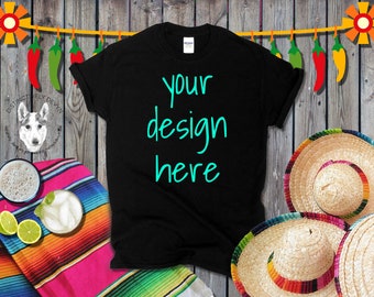 Puede incluir: Camiseta negra con el texto "your design here" escrito en turquesa en la parte delantera. La camiseta está sobre una superficie de madera con una manta mexicana colorida y dos sombreros de paja.