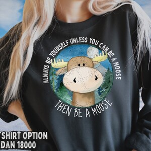 May include: A dark grey sweatshirt featuring a white graphic of a moose in a woodland scene. The text "Always be yourself unless you can be a moose then be a moose" surrounds the image.