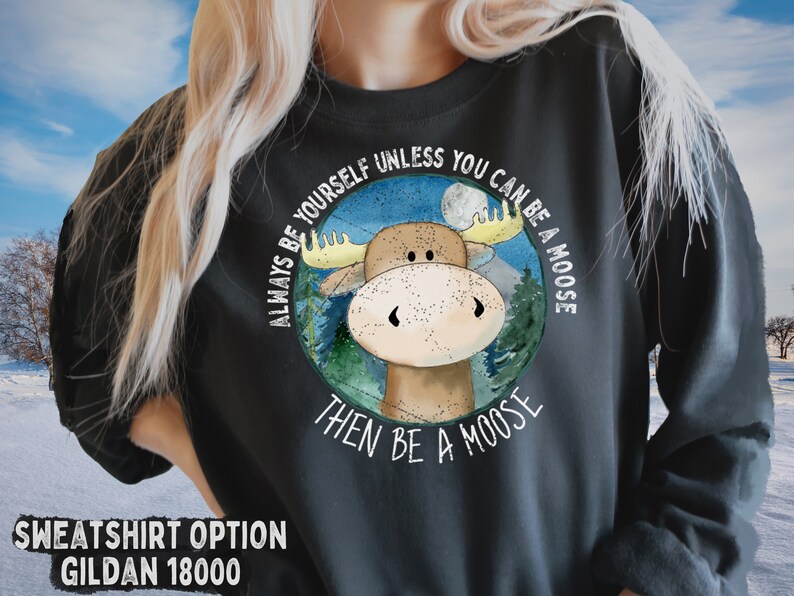 May include: A dark grey sweatshirt featuring a white graphic of a moose in a woodland scene. The text "Always be yourself unless you can be a moose then be a moose" surrounds the image.