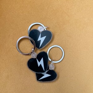 Könnte beinhalten: Drei schwarze herzförmige Schlüsselanhänger mit silbernen Ringen. Jeder Schlüsselanhänger zeigt ein weißes Blitz-Motiv.